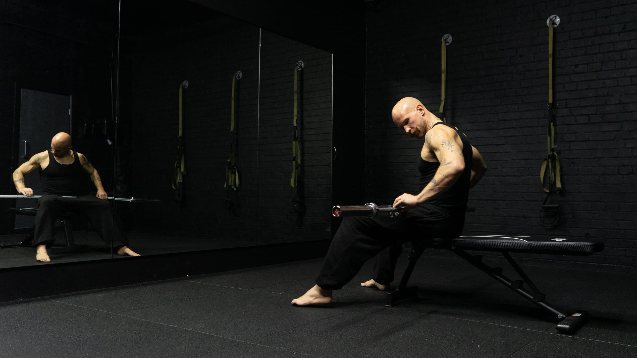 Angus seated on a bench in the gym, working with a bar, his reflection in the mirror behind.